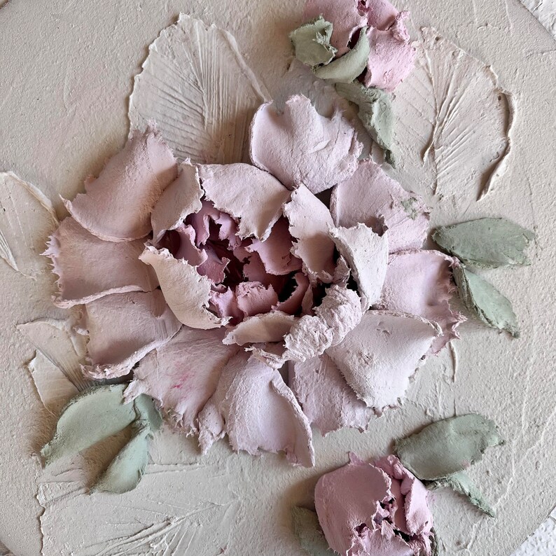 May include: A textured, round, off-white artwork featuring a detailed, raised floral design. The central flower is a soft pink, surrounded by textured petals. Green leaves and smaller pink buds accent the design, creating a three-dimensional effect.