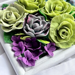 May include: A framed floral arrangement featuring sculpted flowers in shades of green, purple, and gray. The arrangement includes roses, succulents, and small purple flowers, all set within a white wooden frame. The flowers have detailed textures.