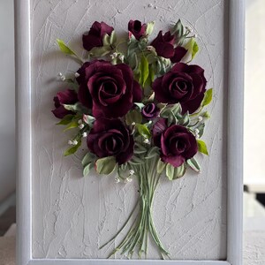 May include: A framed floral artwork featuring a bouquet of deep burgundy roses with green leaves and small white accents. The roses are the focal point, set against a textured, light gray background within a white frame. The artwork has a three-dimensional appearance.