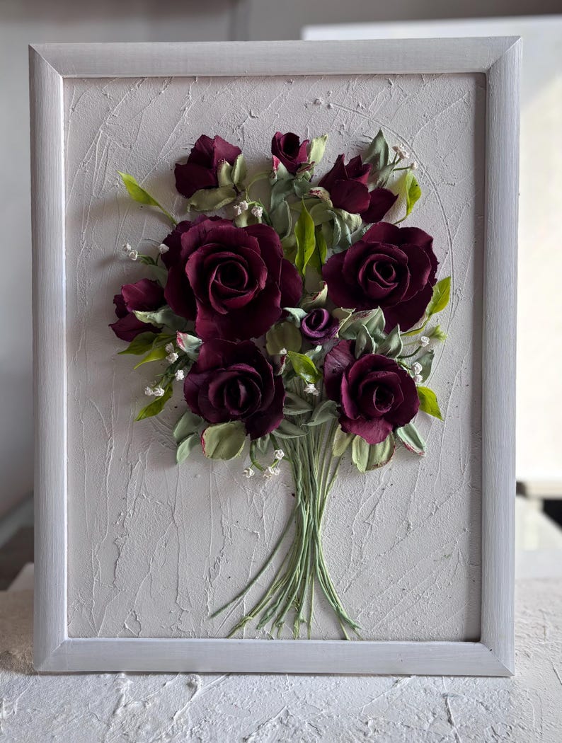 May include: A framed floral artwork featuring a bouquet of deep burgundy roses with green leaves and small white accents. The roses are the focal point, set against a textured, light gray background within a white frame. The artwork has a three-dimensional appearance.