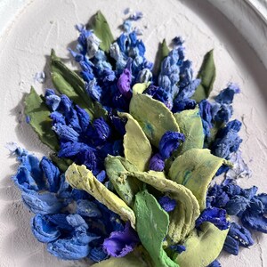 May include: A close-up of a framed painting depicting a bouquet of blue and purple flowers with green leaves. The flowers are painted in a realistic style and appear to be made of textured fabric.