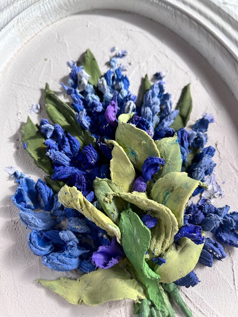May include: A close-up of a framed painting depicting a bouquet of blue and purple flowers with green leaves. The flowers are painted in a realistic style and appear to be made of textured fabric.