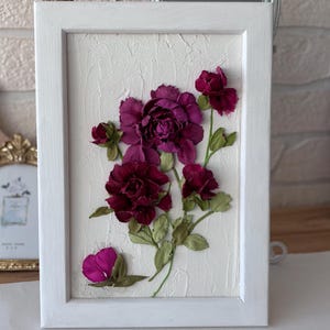 May include: A framed floral artwork featuring deep burgundy peonies and buds with green leaves against a textured white background. The flowers are arranged in a bouquet within a white wooden frame, creating a three-dimensional effect.