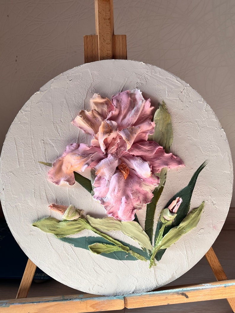K&ouml;nnte beinhalten: Ein rundes Leinwandbild mit einer rosa Iris-Blume mit gr&uuml;nen Bl&auml;ttern und Knospen. Die Blume ist in einem realistischen Stil mit strukturierter Farbe gemalt.