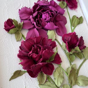 May include: A textured white canvas features a relief of burgundy and purple flowers with green stems and leaves. The flowers are in various stages of bloom, with a large, fully open flower dominating the center. The artwork is framed in white.