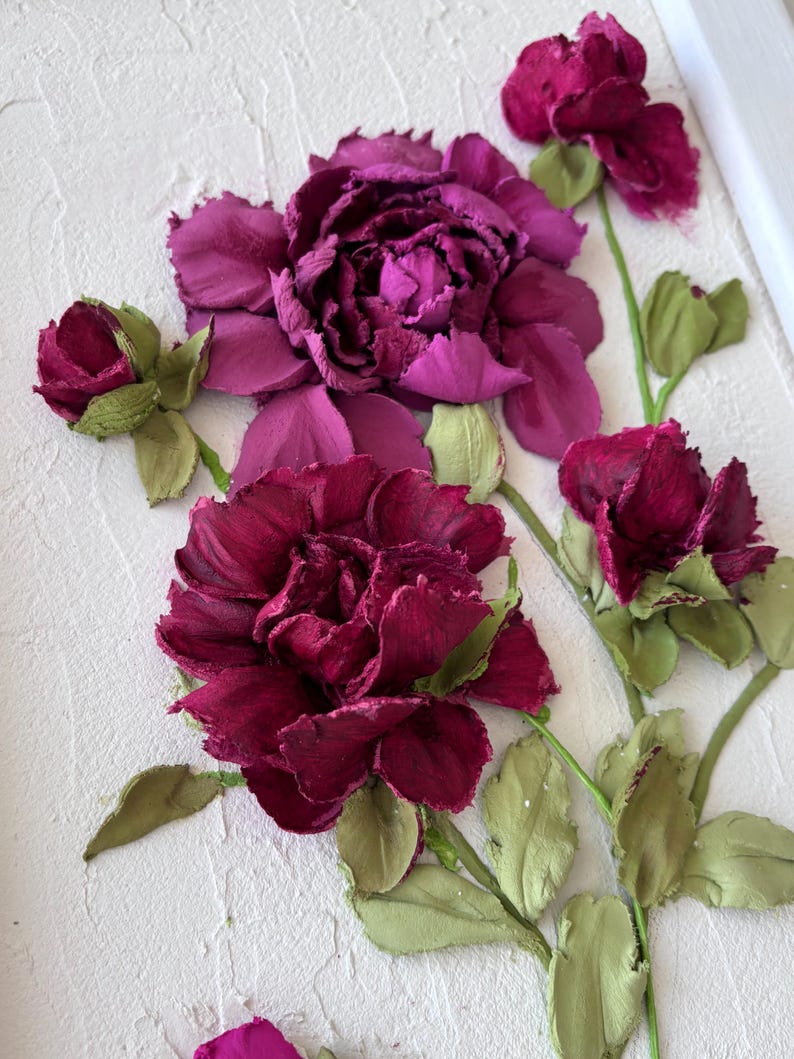 May include: A textured white canvas features a relief of burgundy and purple flowers with green stems and leaves. The flowers are in various stages of bloom, with a large, fully open flower dominating the center. The artwork is framed in white.