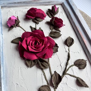 May include: A framed 3D wall art piece featuring a cluster of pink and burgundy roses sculpted from clay. The roses are arranged on a textured white background and are set within a light blue frame.