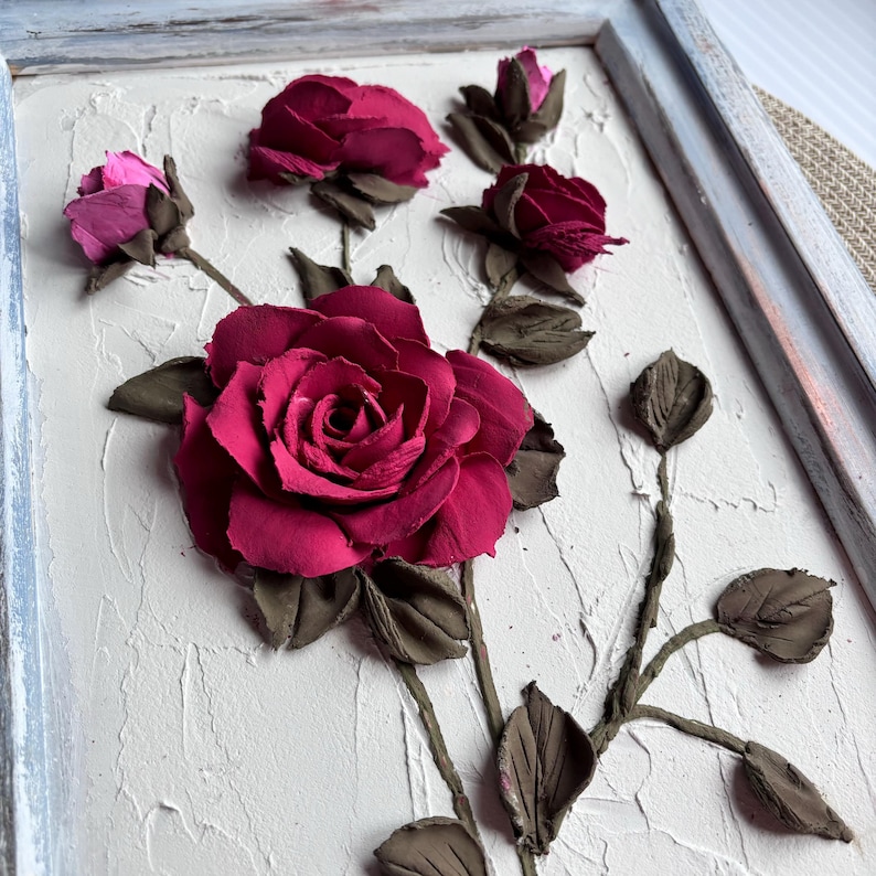 May include: A framed 3D wall art piece featuring a cluster of pink and burgundy roses sculpted from clay. The roses are arranged on a textured white background and are set within a light blue frame.