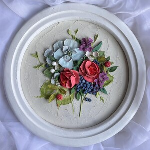 May include: A circular, white-framed floral relief sculpture. The artwork depicts a bouquet of red roses, blue and purple berries, and light blue and purple flowers. Green leaves and white berries complete the design, set against a textured, off-white background.