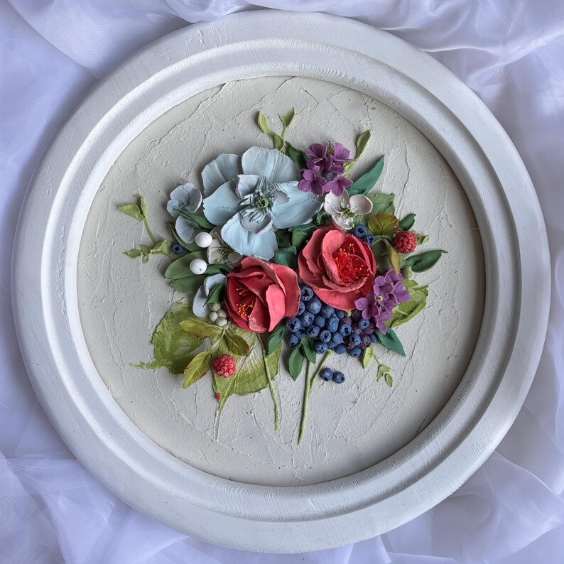 May include: A circular, white-framed floral relief sculpture. The artwork depicts a bouquet of red roses, blue and purple berries, and light blue and purple flowers. Green leaves and white berries complete the design, set against a textured, off-white background.