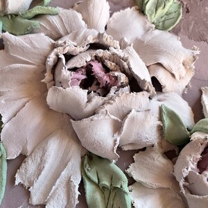 May include: A close-up of a white textured flower with green leaves. The flower is made of a textured material and has a 3D effect.