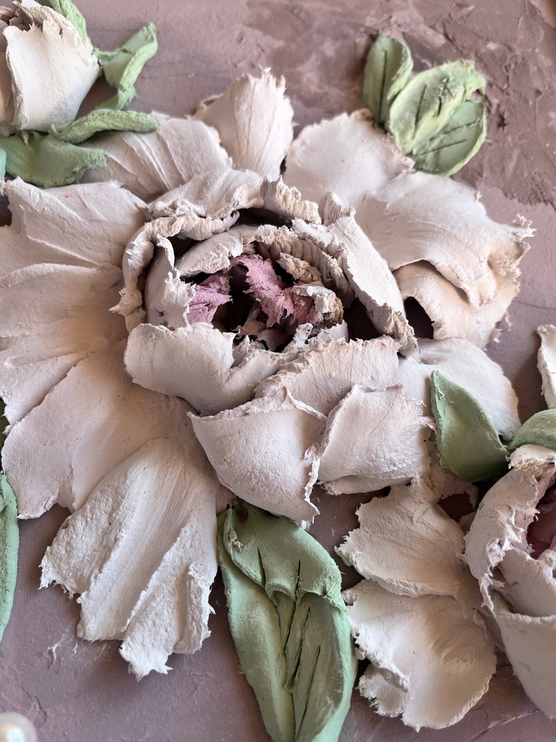 May include: A close-up of a white textured flower with green leaves. The flower is made of a textured material and has a 3D effect.