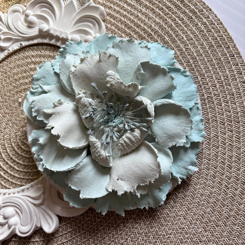 以下が含まれることがあります： テクスチャがあり、しわのある大きな薄い青と白の紙の花。花は画像の中央にあり、主な焦点です。