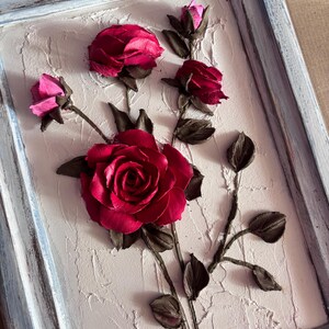 May include: A framed artwork featuring a 3D sculpture of red roses with brown stems and leaves on a textured white background.