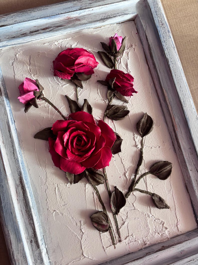 May include: A framed artwork featuring a 3D sculpture of red roses with brown stems and leaves on a textured white background.