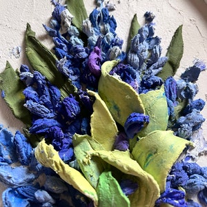 May include: A close-up of a textured painting depicting a bouquet of blue and purple flowers with green leaves. The flowers are painted in a realistic style and have a three-dimensional appearance.