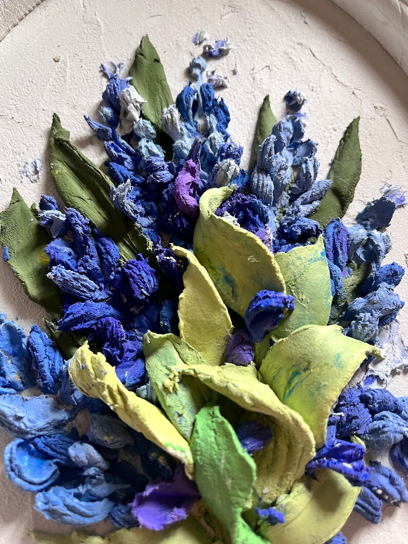 May include: A close-up of a textured painting depicting a bouquet of blue and purple flowers with green leaves. The flowers are painted in a realistic style and have a three-dimensional appearance.