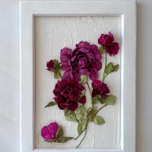 May include: A framed floral artwork featuring burgundy and magenta flowers with green leaves. The flowers are arranged on a textured white background within a white wooden frame. The artwork is a decorative piece.