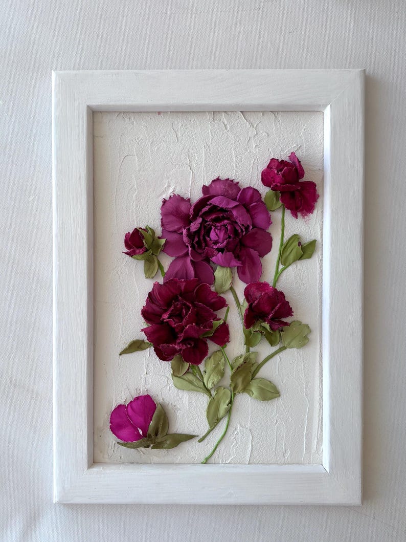 May include: A framed floral artwork featuring burgundy and magenta flowers with green leaves. The flowers are arranged on a textured white background within a white wooden frame. The artwork is a decorative piece.