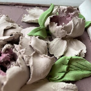 May include: A close-up of a 3D floral wall art piece featuring white and pink peonies with green leaves. The flowers are textured and appear to be made of clay or plaster.