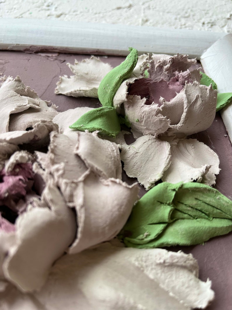 May include: A close-up of a 3D floral wall art piece featuring white and pink peonies with green leaves. The flowers are textured and appear to be made of clay or plaster.