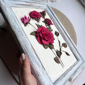 May include: A framed artwork featuring a 3D floral design with pink and red roses on a white background. The frame is a light blue color with a distressed finish.