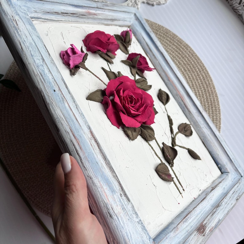 May include: A framed artwork featuring a 3D floral design with pink and red roses on a white background. The frame is a light blue color with a distressed finish.