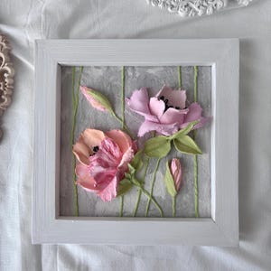 May include: A framed floral artwork featuring two dimensional flowers in shades of pink and peach. The flowers are set against a gray background with green vertical lines. The frame is white.