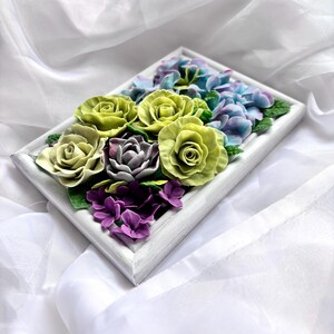 May include: A rectangular white framed floral arrangement featuring sculpted flowers. The arrangement includes green roses, purple hydrangeas, and other flowers in shades of purple and blue. The flowers are set against a white background.