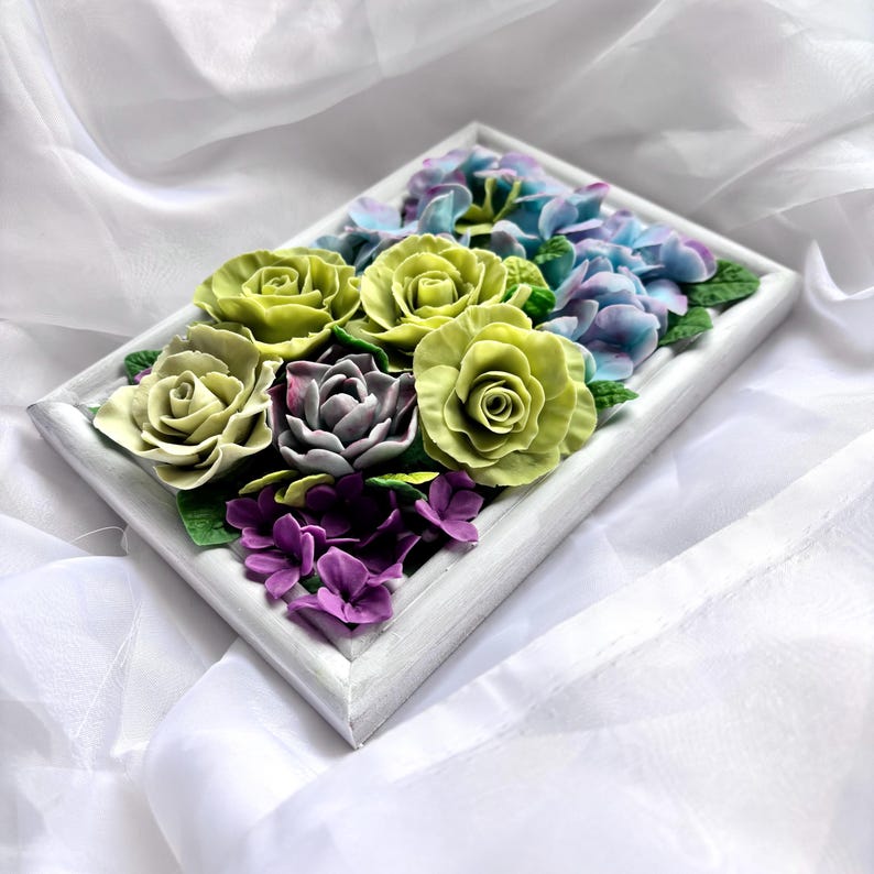 May include: A rectangular white framed floral arrangement featuring sculpted flowers. The arrangement includes green roses, purple hydrangeas, and other flowers in shades of purple and blue. The flowers are set against a white background.