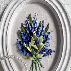 May include: A white oval frame with a textured surface, featuring a 3D floral painting of a bouquet of blue and green flowers. The flowers are arranged in a cluster and appear to be made of fabric or felt.