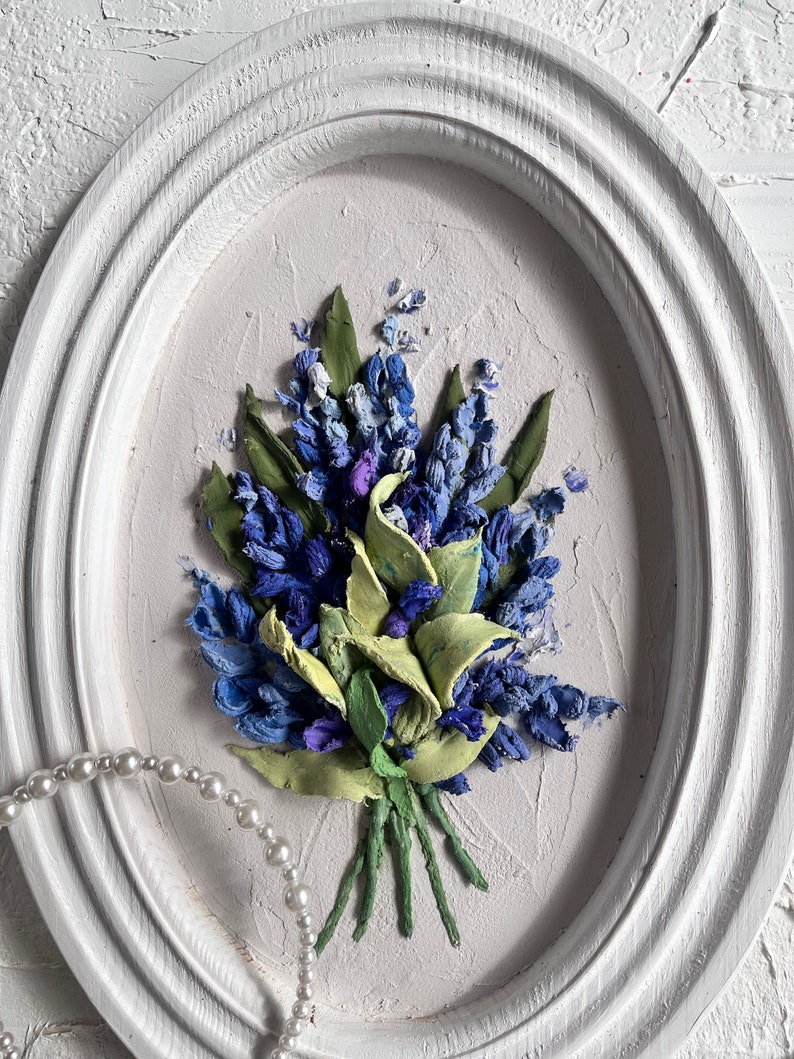 May include: A white oval frame with a textured surface, featuring a 3D floral painting of a bouquet of blue and green flowers. The flowers are arranged in a cluster and appear to be made of fabric or felt.