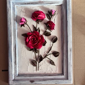 May include: A framed artwork featuring a white background with a textured surface. The artwork depicts a cluster of red roses with pink buds and brown stems.