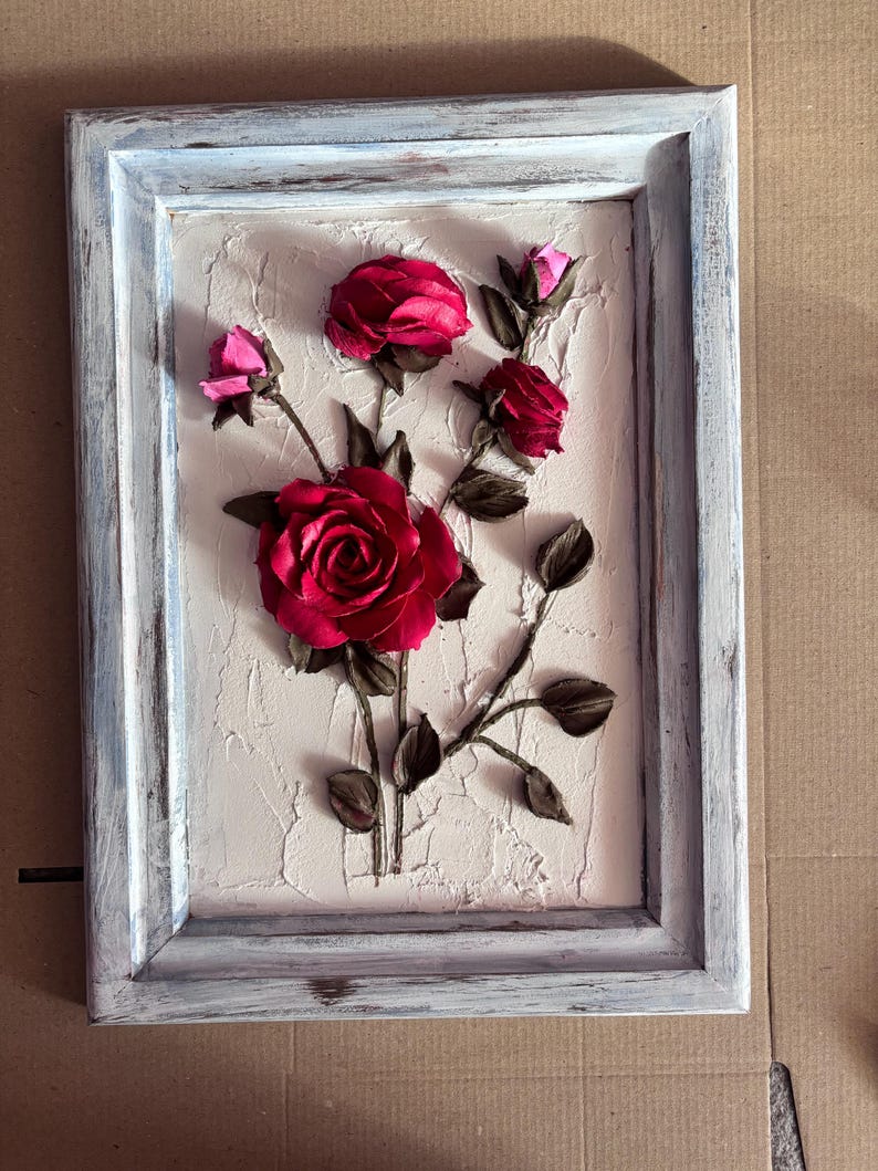May include: A framed artwork featuring a white background with a textured surface. The artwork depicts a cluster of red roses with pink buds and brown stems.