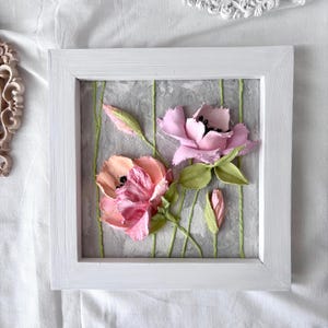 May include: A square white framed artwork featuring textured, three-dimensional flowers. The flowers are in shades of pink and peach with green leaves and stems against a gray background. The artwork is approximately 8 inches square.