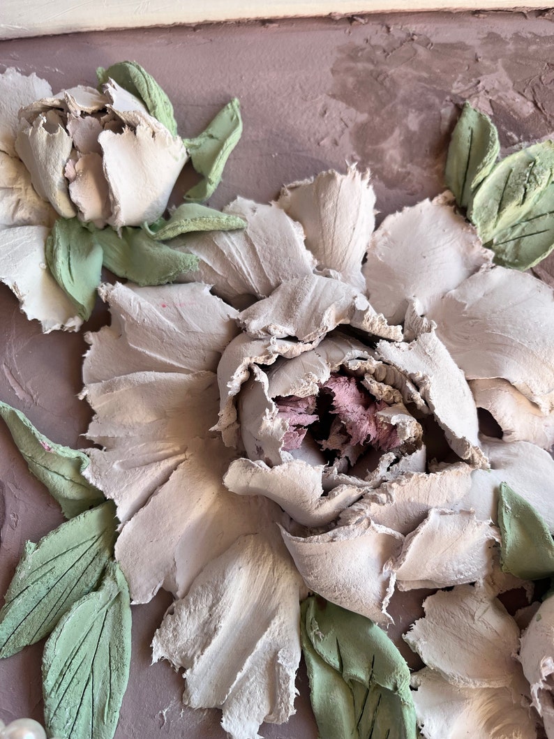 May include: Close-up of a white, textured, 3D flower with green leaves. The flower is made of a material that resembles clay or plaster.