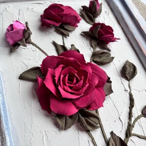 May include: A close-up of a 3D wall art piece featuring a cluster of pink and red roses sculpted from clay. The roses are arranged on a textured white background.