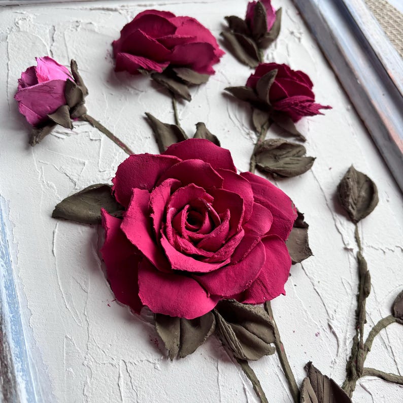 May include: A close-up of a 3D wall art piece featuring a cluster of pink and red roses sculpted from clay. The roses are arranged on a textured white background.