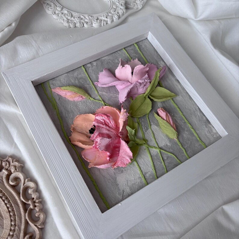 May include: A framed floral artwork featuring textured, three-dimensional flowers in shades of pink and peach. The flowers are set against a gray background with green stems, enclosed in a white wooden frame. The artwork evokes a sense of spring.