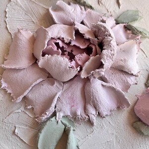 May include: A close-up of a textured, light pink floral relief sculpture. The flower has multiple layers of petals in varying shades of pink, with green leaves. The background is a light beige color.