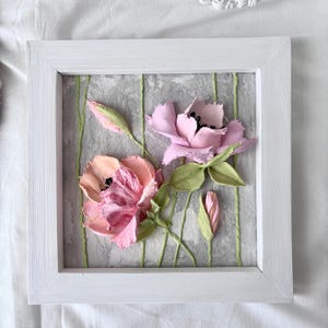May include: A framed floral artwork featuring two textured, pink flowers with black centers and green leaves. The flowers are set against a gray background with vertical green lines, all within a white wooden frame. The artwork has a 3D effect.