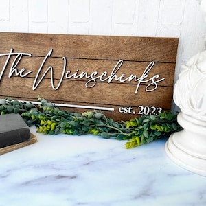 May include: A wooden sign with the text "The Weinschenks est. 2023" in white lettering. The sign has a rustic brown finish and is made of wood planks.