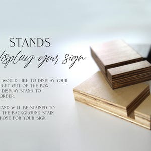 May include: Wooden display stands for signs. The stands are made of plywood and are stained to match the background stain of the sign. "STANDS To display your sign" is written on the white background.
