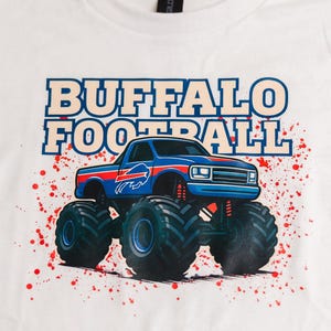 K&ouml;nnte beinhalten: Wei&szlig;es T-Shirt mit einem blauen Monster-Truck-Design, dem Buffalo Bills-Logo und den Worten "BUFFALO FOOTBALL". Das Design enth&auml;lt rote Farbspritzer. Ein l&auml;ssiges Kleidungsst&uuml;ck.