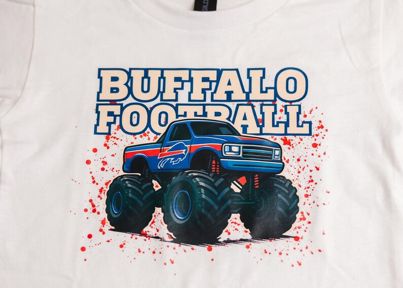 K&ouml;nnte beinhalten: Wei&szlig;es T-Shirt mit einem blauen Monster-Truck-Design, dem Buffalo Bills-Logo und den Worten "BUFFALO FOOTBALL". Das Design enth&auml;lt rote Farbspritzer. Ein l&auml;ssiges Kleidungsst&uuml;ck.