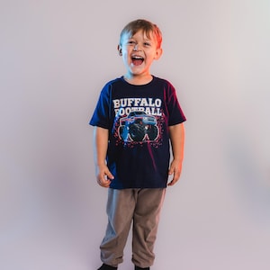 K&ouml;nnte beinhalten: Ein Kind tr&auml;gt ein marineblaues T-Shirt mit der Aufschrift "BUFFALO FOOTBALL" und einer Grafik eines Monstertrucks. Das Kind tr&auml;gt auch eine graue Hose und schwarze Socken. Das Kind l&auml;chelt und lacht.