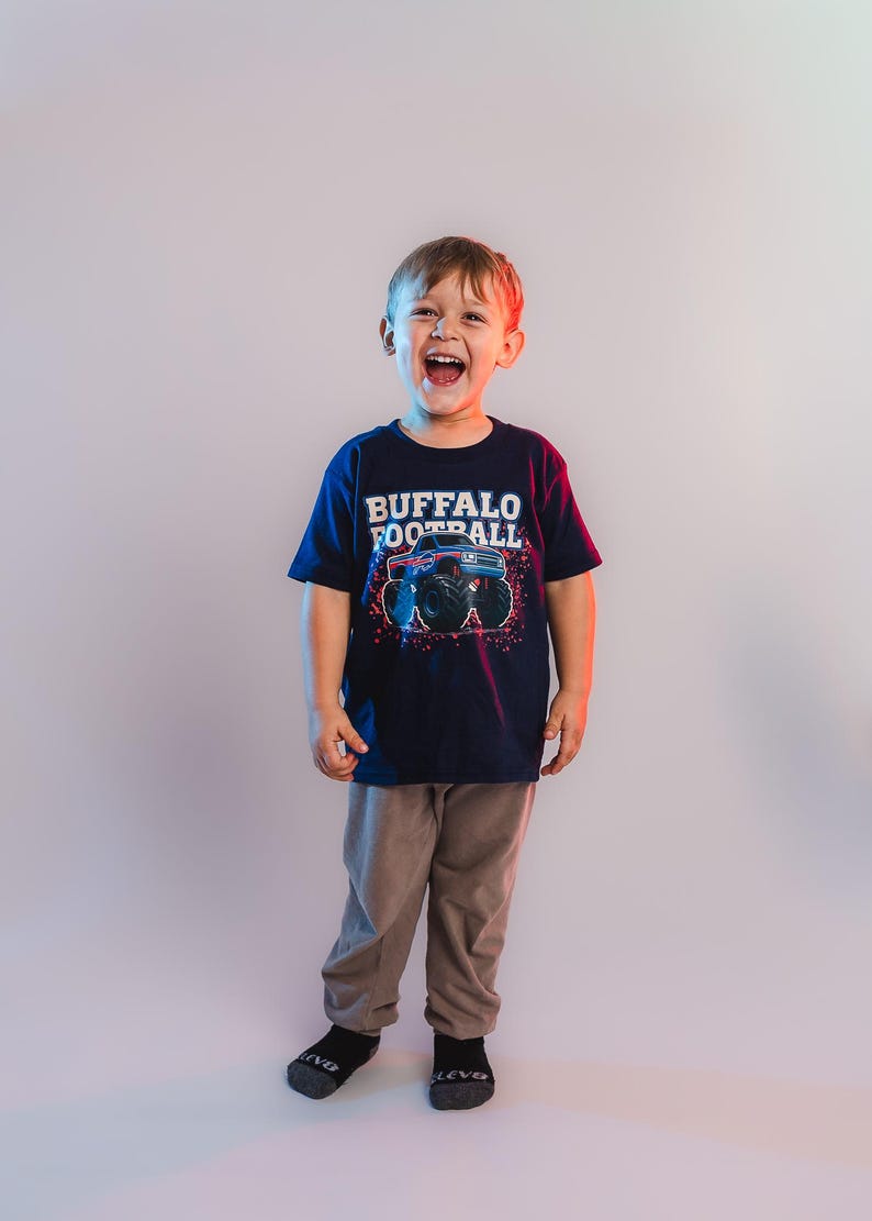 K&ouml;nnte beinhalten: Ein Kind tr&auml;gt ein marineblaues T-Shirt mit der Aufschrift "BUFFALO FOOTBALL" und einer Grafik eines Monstertrucks. Das Kind tr&auml;gt auch eine graue Hose und schwarze Socken. Das Kind l&auml;chelt und lacht.