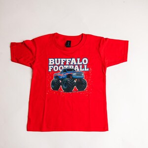 K&ouml;nnte beinhalten: Ein rotes T-Shirt mit einer Grafik eines blauen Monstertrucks mit gro&szlig;en schwarzen Reifen. Der Text "BUFFALO FOOTBALL" ist in Wei&szlig; und Blau &uuml;ber dem Truck aufgedruckt. Das Shirt ist aus einem weichen Material.