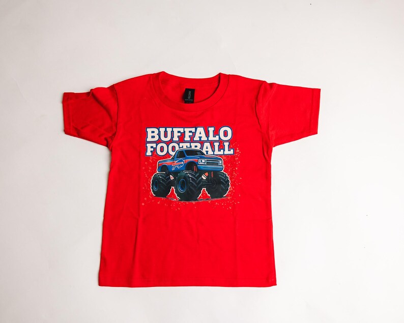 K&ouml;nnte beinhalten: Ein rotes T-Shirt mit einer Grafik eines blauen Monstertrucks mit gro&szlig;en schwarzen Reifen. Der Text "BUFFALO FOOTBALL" ist in Wei&szlig; und Blau &uuml;ber dem Truck aufgedruckt. Das Shirt ist aus einem weichen Material.