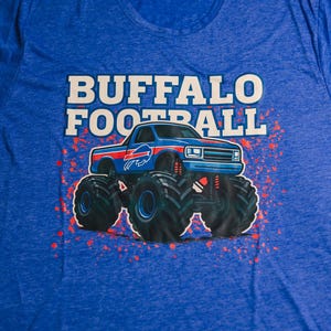K&ouml;nnte beinhalten: Blaues T-Shirt mit dem Schriftzug "BUFFALO FOOTBALL" in wei&szlig;en Blockbuchstaben &uuml;ber einer Monster-Truck-Grafik. Der Truck ist blau mit roten und wei&szlig;en Akzenten und gro&szlig;en schwarzen Reifen. Rote, spritzerartige Akzente umgeben den Truck.